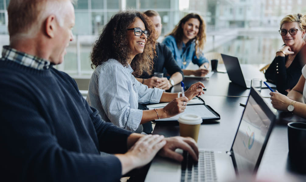 six office workers smile at a conference table meeting with coffee, notebooks, and laptops